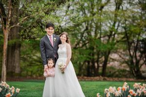A bride and groom pose with their young daughter in a lush garden setting with blooming daffodils and spring trees.
