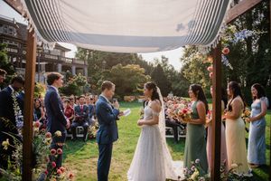 A romantic outdoor wedding ceremony takes place under a wooden pergola with rolling hills in the background at sunset.