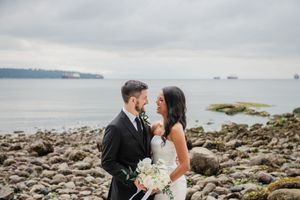 A couple shares a romantic moment on a rocky beach coastline with gray skies, holding a white floral bouquet.