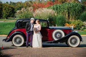 Couple poses beside vintage red and black classic car with spoked wheels in garden setting.