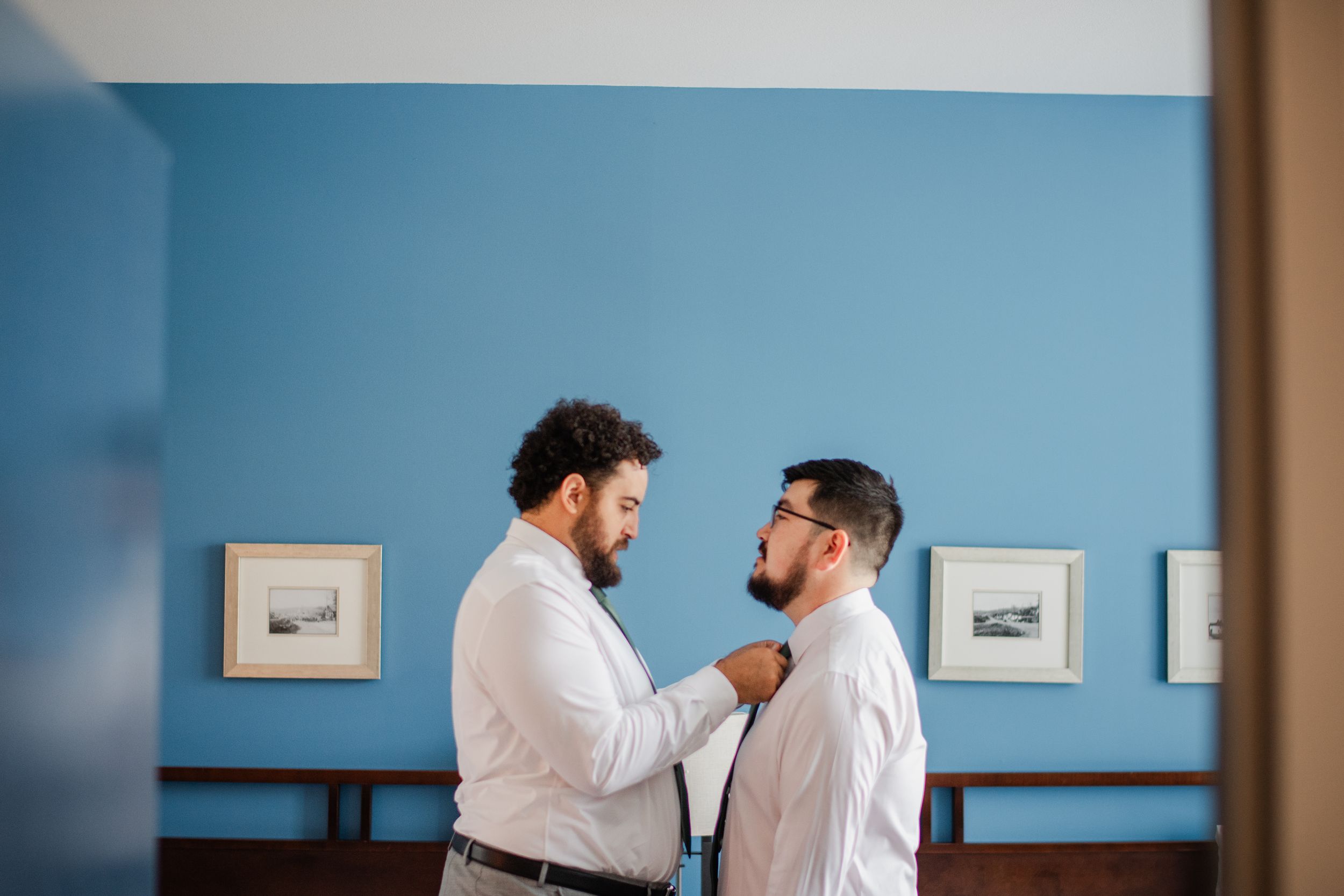 Two groomsmen in white shirts help each other with a tie while getting ready in a blue-walled room.