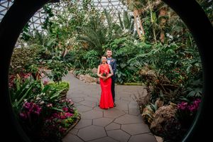 A figure in a vibrant red dress stands in a lush greenhouse conservatory surrounded by tropical plants and flowers.
