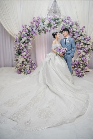 Elegant wedding scene with white and purple floral arch and long trailing lace wedding dress.