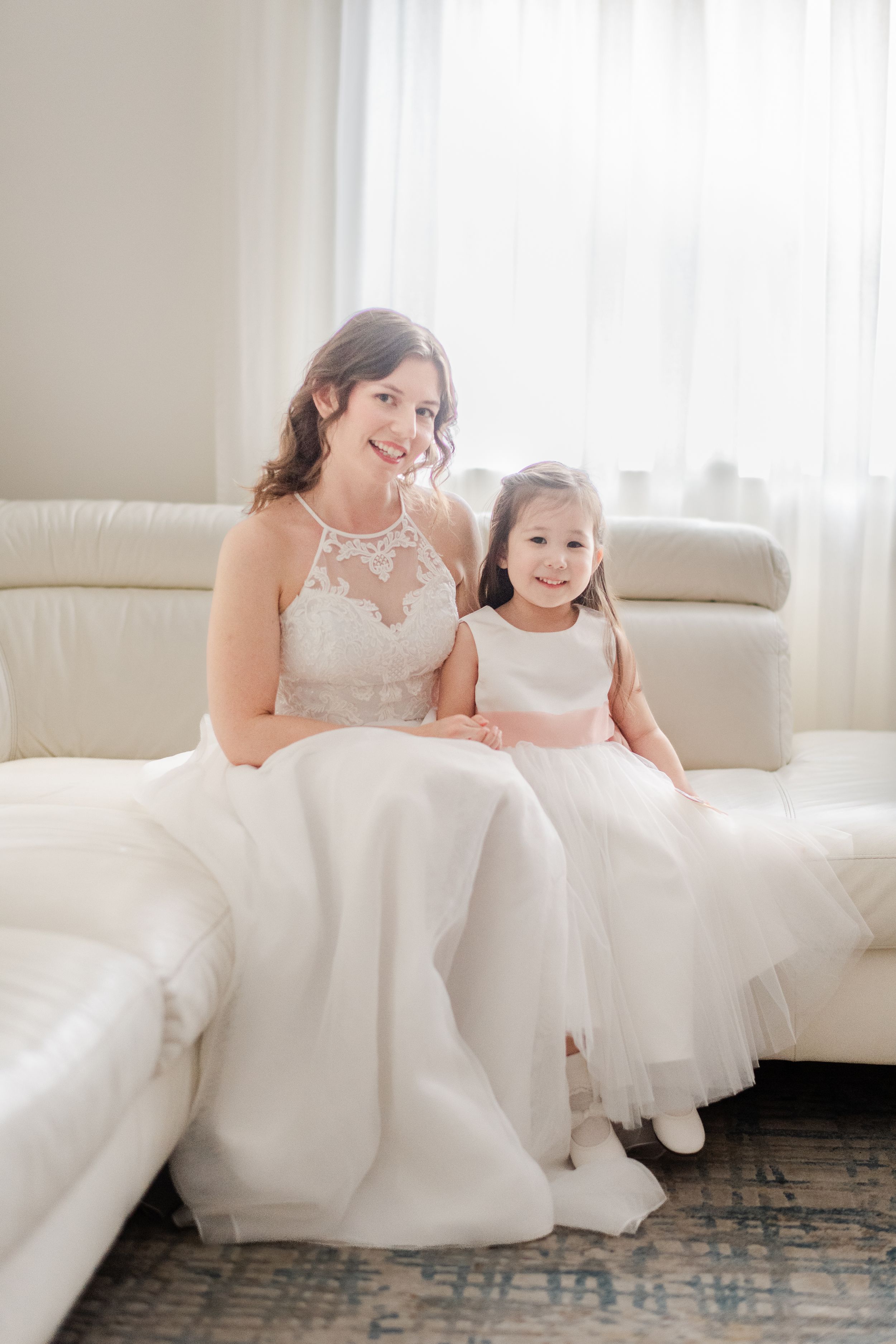 Bride in lace gown sits with flower girl in white dress on ivory sofa in bright elegant room.