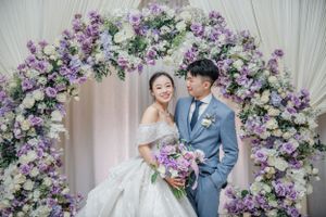 Wedding couple posing under purple and white floral arch with elegant decor.