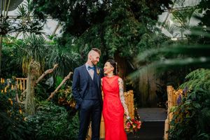 Two people stand together on a stone path surrounded by tropical plants and foliage in a greenhouse.