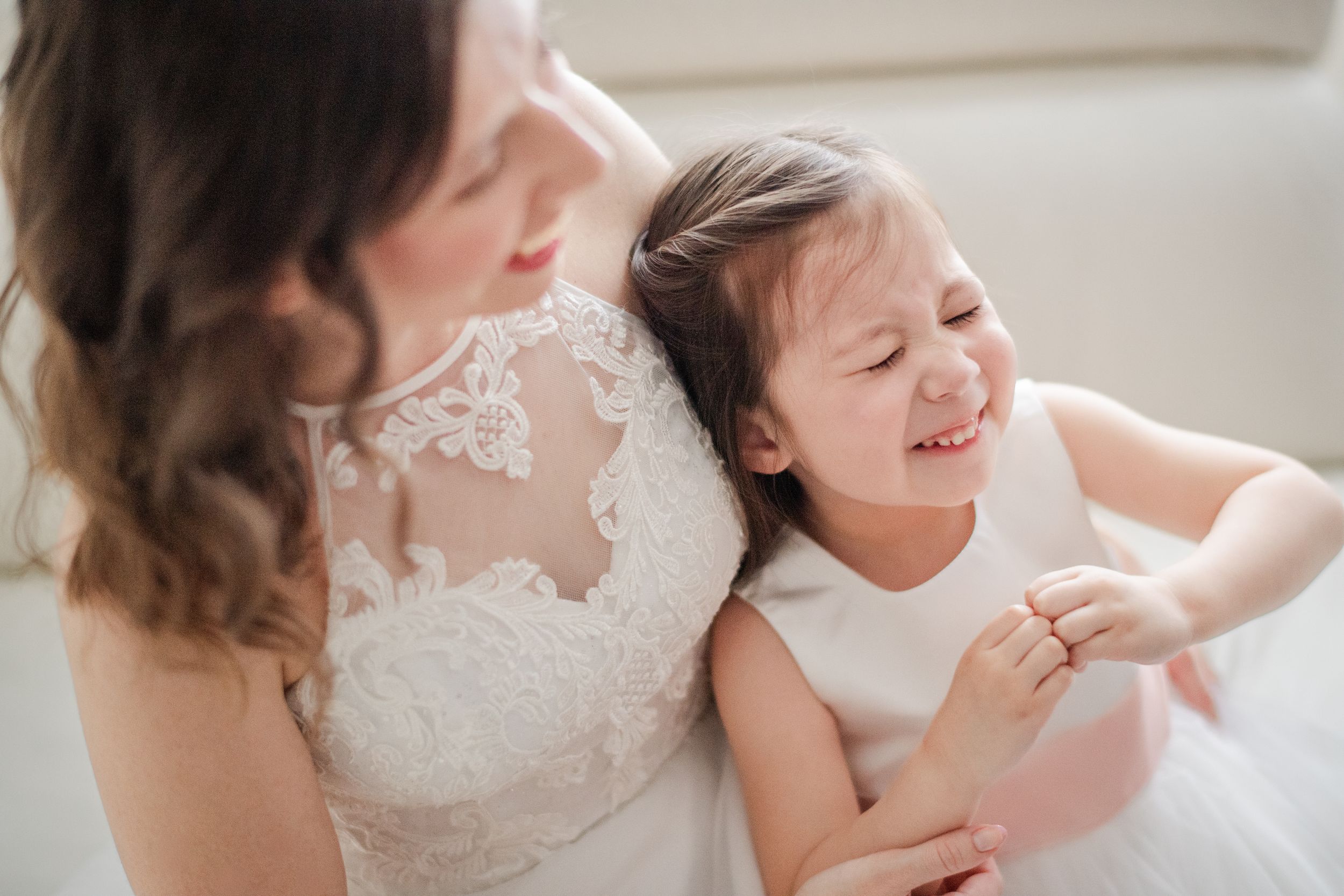 A bride in lace wedding gown shares a joyful moment with a laughing little girl in white dress.