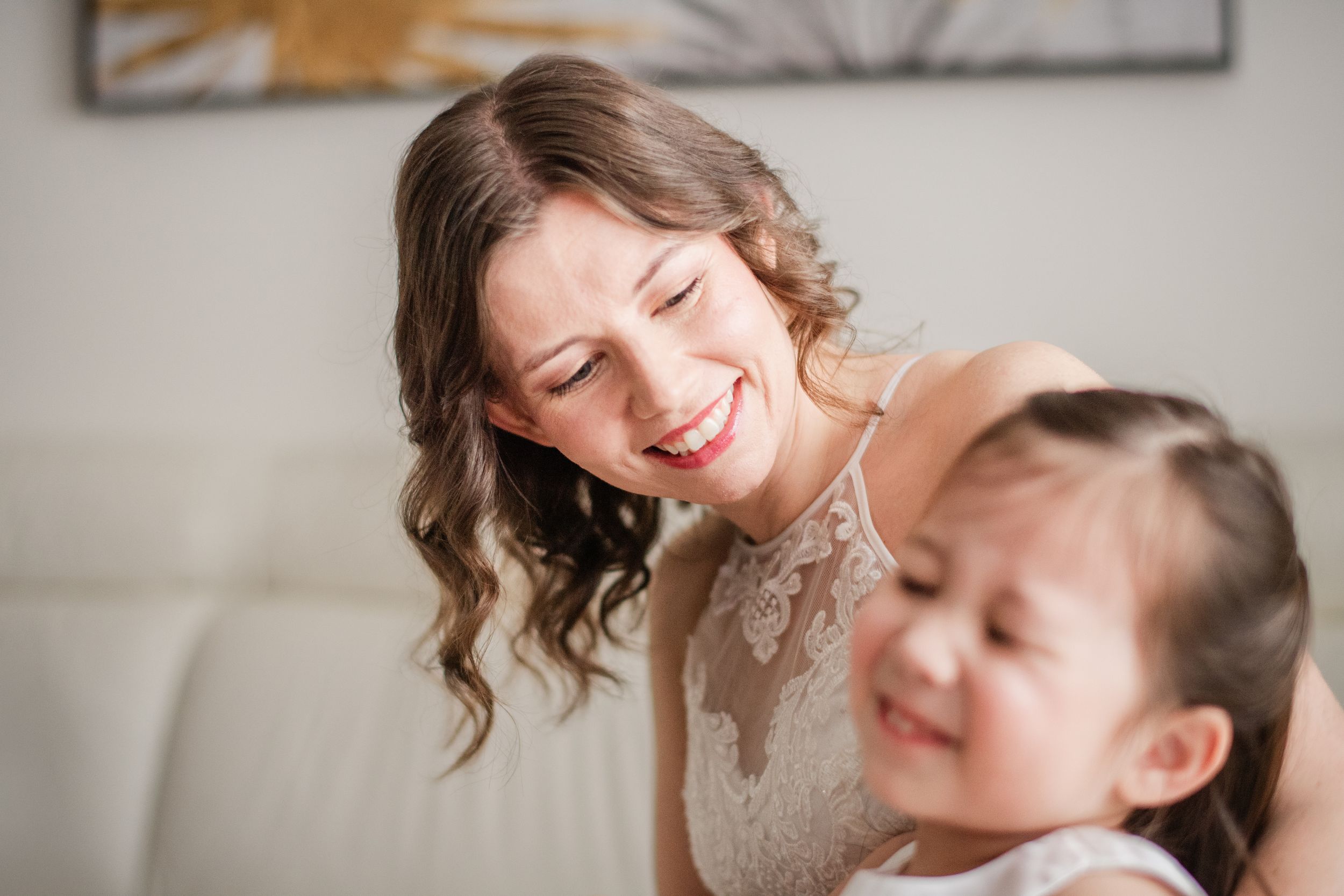 A smiling bride in lace dress laughing with a young flower girl on a white sofa during wedding preparations.