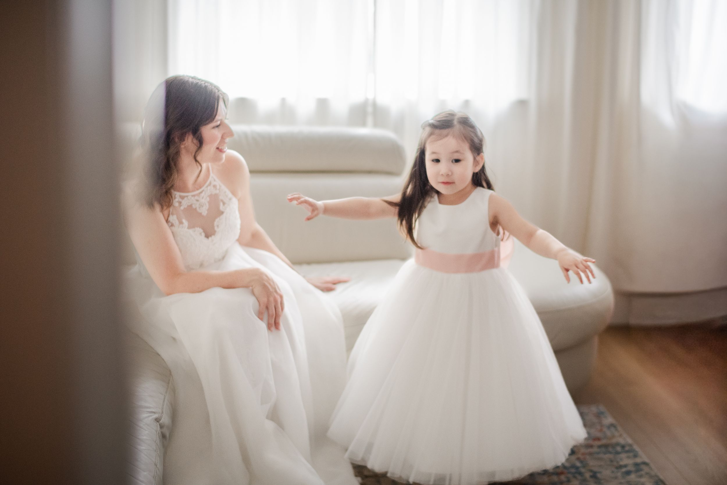 A bride in a lace gown sits beside a young flower girl in a white tulle dress with pink sash in a bright room.