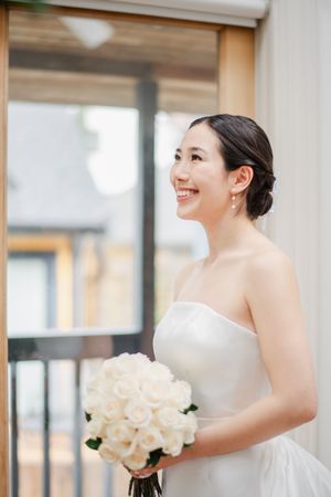 A bride wearing a white strapless wedding dress smiles while holding a bouquet of cream roses in an indoor setting.