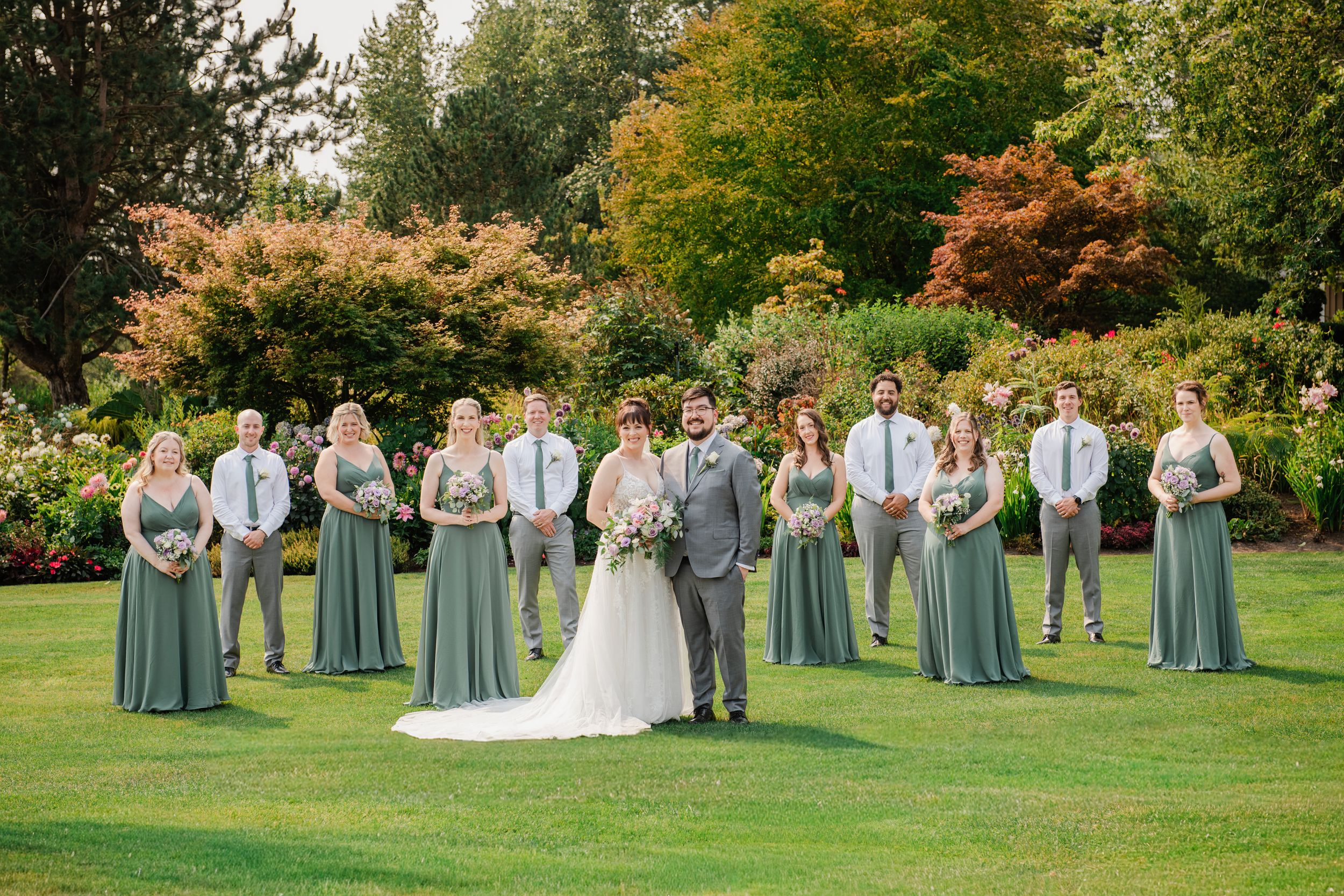 Wedding party in sage green attire posed on lush lawn with colorful autumn trees backdrop.