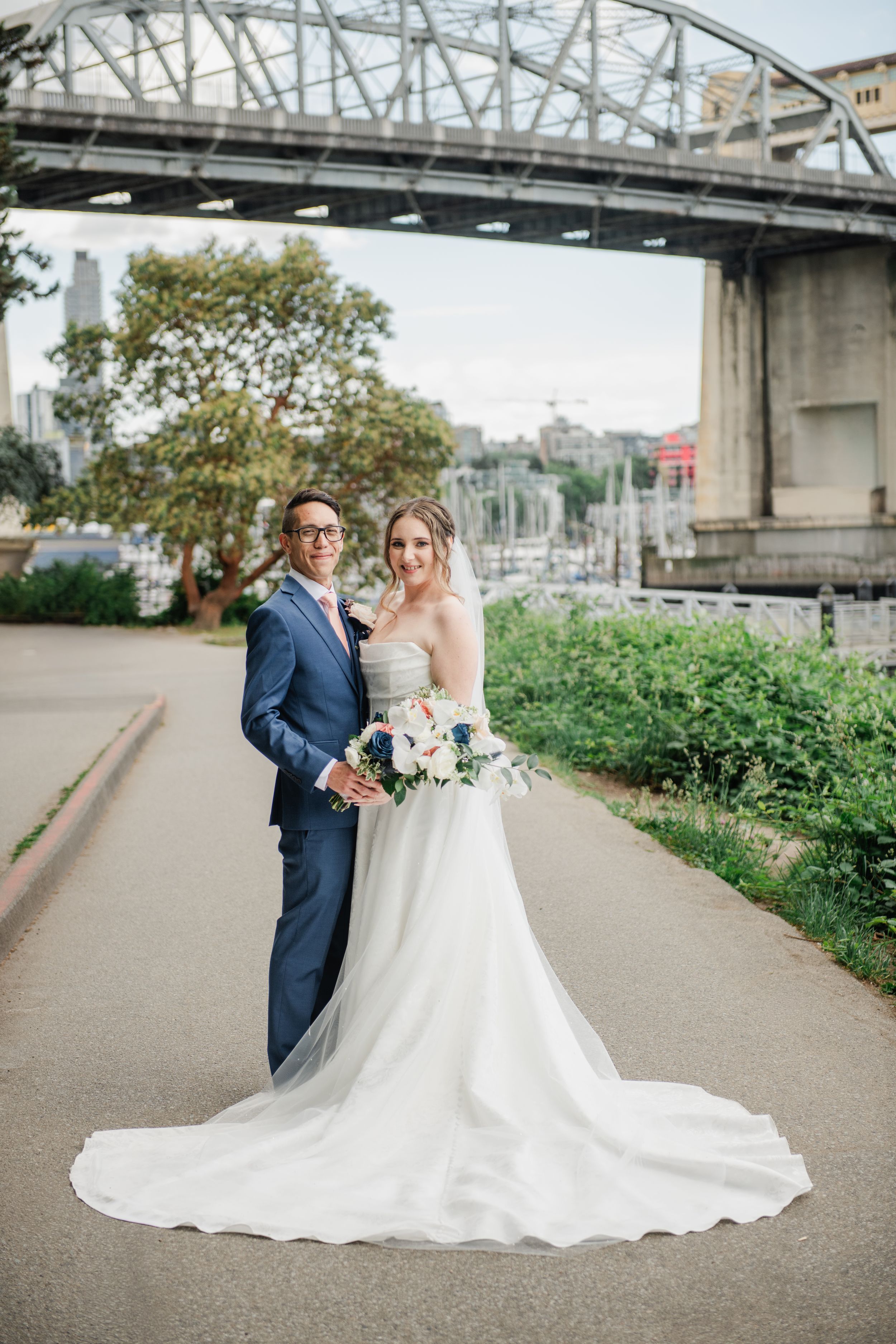 Bride and groom posing near a bridge waterfront, bride in white gown with long train holding floral bouquet.