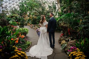 A couple shares a romantic moment in a lush greenhouse conservatory filled with tropical plants and colorful flowers.