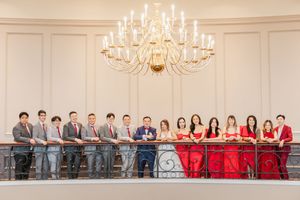 Wedding party stands in elegant ballroom with chandeliers and red decor against cream colored walls.