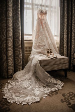 A delicate lace wedding dress with long train hangs by a window with ornate curtains.