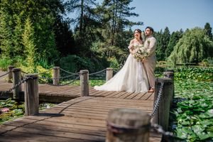 A newlywed couple stands on a wooden bridge over lily pads in a serene garden with evergreen trees in the background.