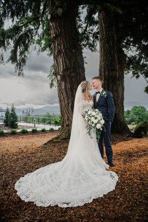 A wedding portrait beneath towering evergreen trees with a dramatic cloudy sky backdrop.