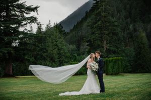 A couple embraces on a grassy lawn with mountains and evergreen forest in the background.
