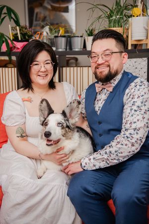 A smiling couple in wedding attire sits together holding a fluffy Corgi dog, surrounded by plants indoors.