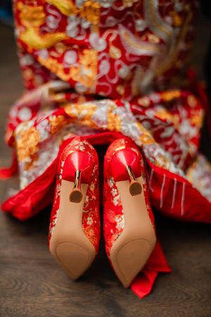 Close up of traditional red bridal shoes with ornate embellishments displayed on a dark wooden floor.