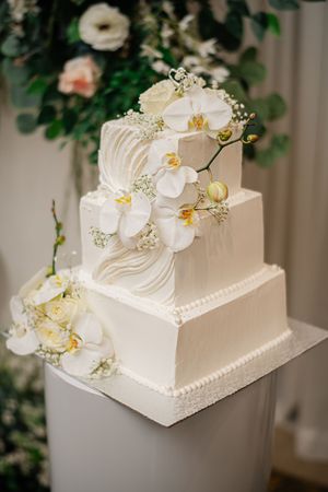 Elegant three-tier square white wedding cake adorned with white orchids and baby's breath on a white pedestal.