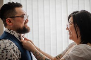 A smiling woman adjusts a bow tie on a bearded man wearing glasses and a blue suit jacket.