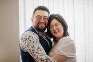 A smiling couple in glasses share a warm embrace, dressed in semi-formal attire against a bright window backdrop.