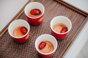Four red and white ceramic cups of tea with red dates on a woven tray.