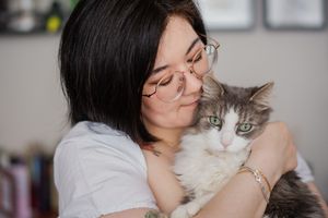 Young woman with glasses hugging a fluffy gray and white cat, sharing a tender moment at home.