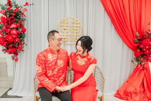 Couple in matching red traditional Chinese outfits sitting together, smiling, with red floral arrangements and draping backdrop.