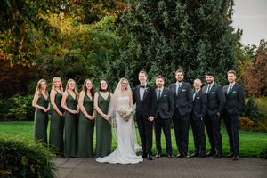 Wedding party stands in formal attire against autumn foliage backdrop in outdoor garden setting.