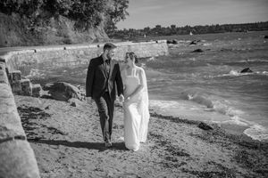 A couple in formal attire walks together along a rocky beach shoreline in black and white photography.