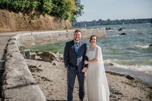 A couple stands together on a rocky beach seawall with waves and coastline in the background.