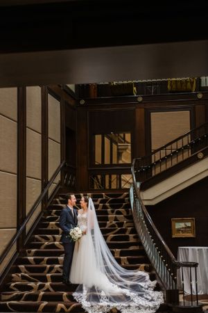A couple poses on an elegant wooden staircase with a flowing lace veil trailing down the steps.