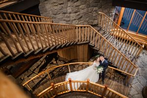 Couple in wedding attire embracing on a grand wooden spiral staircase with stone walls and warm lighting.