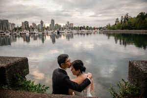 A couple embraces by a serene waterfront with Vancouver's skyline reflected in the calm harbor at sunset.