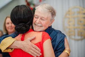Elderly man with white hair smiling warmly while hugging a woman in a red dress at a celebration.