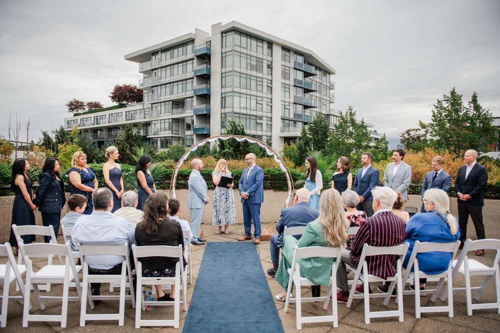 Outdoor wedding ceremony with guests seated in white chairs, couple at floral arch, modern building backdrop.