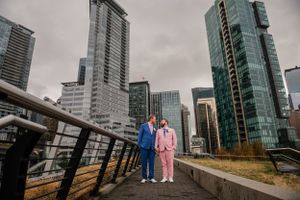 Couple in vibrant blue and pink outfits pose against modern city skyline with glass skyscrapers on cloudy day.