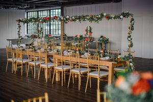 Long banquet table with wooden chairs decorated with hanging greenery garlands and floral arrangements in an industrial venue.