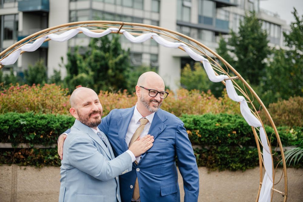 Two grooms in blue suits share a joyful moment under a gold circular arch at an outdoor wedding ceremony.
