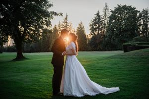 Couple in formal wedding attire walks on lush green lawn at sunset with tall evergreen trees in background.