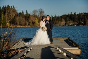 Newlyweds share a moment on a wooden dock overlooking a serene lake and forest.