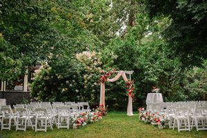 Outdoor wedding ceremony setup with floral arch, white chairs, and lush green trees in a garden setting.