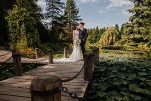 Couple embraces on rustic wooden bridge over lily-covered pond with evergreen trees in background.