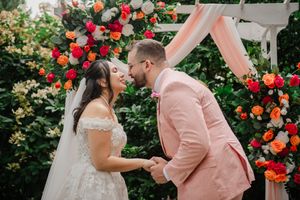 Bride and groom share a kiss at outdoor wedding ceremony with colorful floral arch of red, orange, and pink roses.