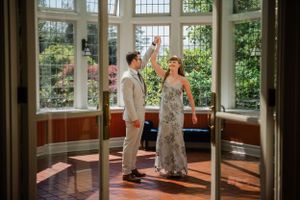Couple in formal wear standing in sunlit conservatory with large windows.