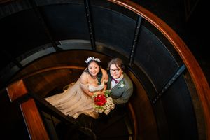Wedding couple walks down wooden spiral staircase with railing in dramatic lighting and dark background.
