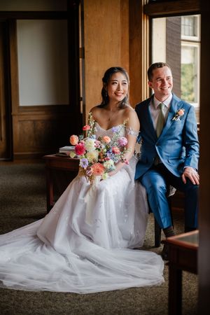 A couple in wedding attire sits together on a wooden bench in a rustic indoor setting with beautiful floral bouquet details.