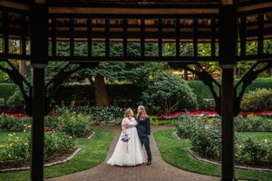 A couple embraces under a wooden pergola surrounded by lush gardens and blooming flowers at a romantic outdoor venue.